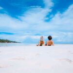 Two young travelers sitting on rustic wooden stools along a white sand beach overlooking a turquoise ocean with a traditional dhow boat, using a curated Zanzibar travel guide for an island escape.