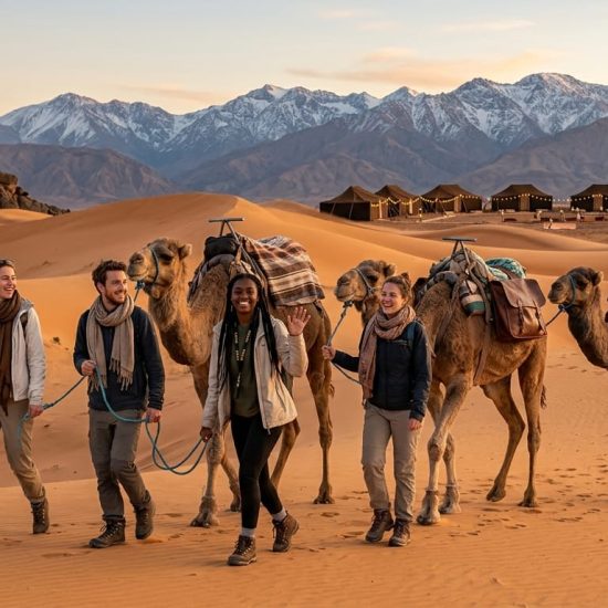 A group of five young travelers from a Kito Afrika Morocco group trip 2026, smiling as they walk with camels through Sahara dunes toward the Atlas Mountains during a golden sunset, designed for the November itinerary guide.